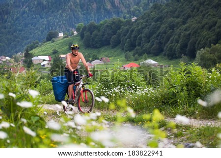 a man riding a mountain bike on a gravel road in a day. background green and rocky mountains, meadows and a village of white flowers in the background. view from the face