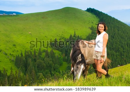 happy girl sitting on a green flower meadow near a wild horse and her calf. background of green hills
