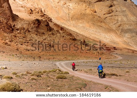 Two cyclists ride on a winding dirt road red pitch in Canyon Morocco Dades in Atlas Mountains, Africa. Background blue sky with clouds. Beautiful World.