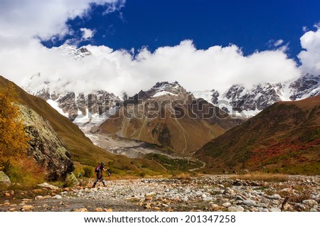 Girl jumps over the river, using trekking poles with a backpack. Autumn landscape. Composition of nature. Mountain River into canyon, valley. Blue clouds.