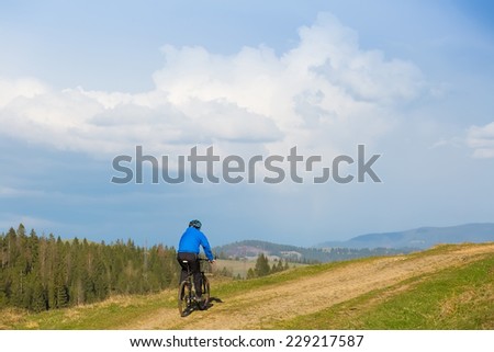 mountain biker on sunny day riding on a winding dirt road in a rural hilly area of green forest against the blue sky with beautiful clouds