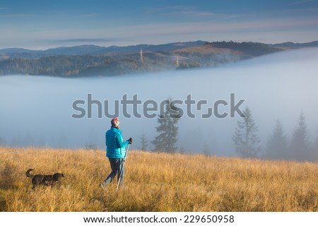 Girl with a dog goes towards the house standing on a mountain with Trekking pole in the morning fog. Landscape composition, background mountains and sunrise.