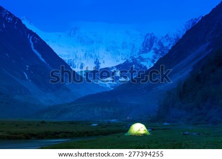 Glowing tent stands on the banks of a mountain stream, amid high mountains and snow-capped peaks. Twilight, night.
