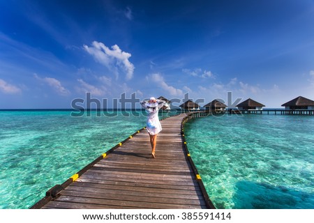 Beautiful girl in white dress and hat standing on wooden planks in the Maldives island on the background of a bungalow on the water and the beauty of the sea with the coral reefs