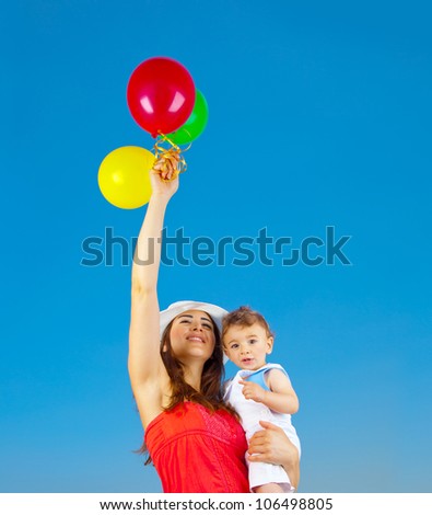 Happy family holding colorful air balloons over blue sky background, mother and adorable child baby boy playing games outdoor, mum and kid having fun in summer nature, hands up, freedom joy concept