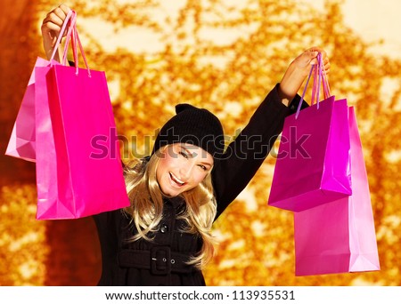 Image of happy cute girl with pink shopping bag, cheerful young lady holding paper presents bags in fall park, smiling blond woman raised up hands over golden autumn background, spending money concept
