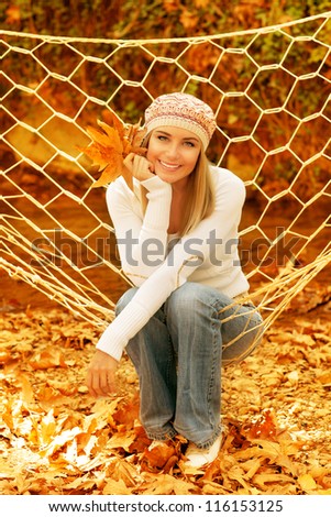 Picture of cute female swinging in hammock in autumnal park, closeup portrait of pretty woman relaxed on backyard, attractive blond young lady holding golden dry tree leaves in hand