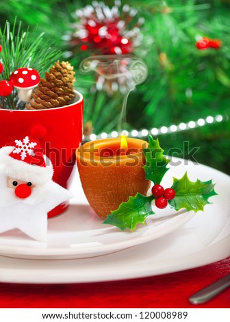 Image of romantic Christmastime dinner with warm candle light, red coffee cup decorated with Santa Claus star and fir cone, beautiful white dinnerware on Christmas tree background, New Year party