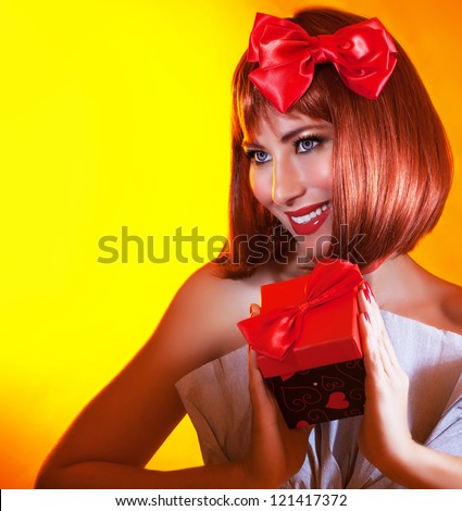 Picture of pretty woman with red bow in hair holding gift box in hands, gorgeous female with red hair isolated on yellow background, attractive girl wearing stylish festive dress, New Year party