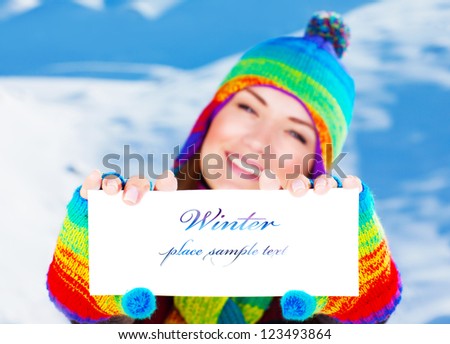 Picture of cute happy woman holding paper card with text in hands, pretty cheerful female having fun outdoors in wintertime, wearing stylish colorful winter hat and gloves, shallow depth of field