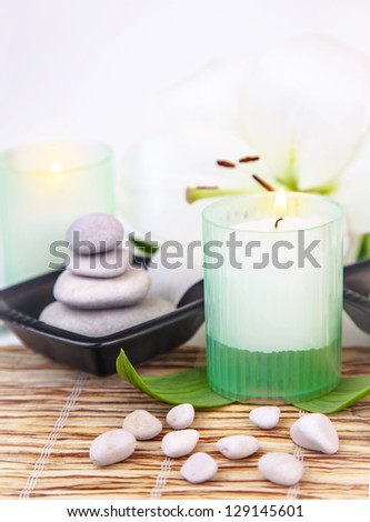 Image of pebble stones in black plate, white lily flower, fresh green leaves, candle on bamboo table in spa salon, natural cosmetics, relaxation in dayspa, meditation and harmony concept