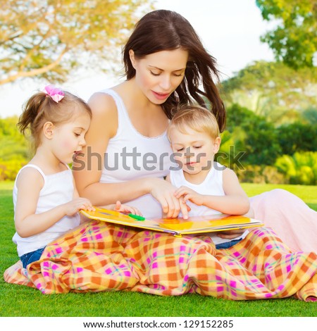 Image Of Cute Young Female With Two Little Children Read Book Outdoors, Brother And Sister With Mother Learning In The Park In Spring Time, Preschool Education, Day Care, Happy Family Concept