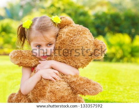 Little cute sad girl holding in hands brown teddy bear, upset child spending time outdoors in spring time