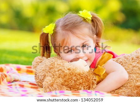 Cute smiling baby girl hugging soft bear toy, sweet kid having fun outdoors in day care, laying down on green grass in spring