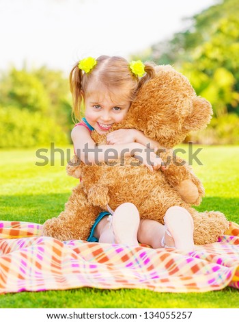 Cute little happy girl hugging big brown teddy bear and sitting down on green grass meadow, spring season