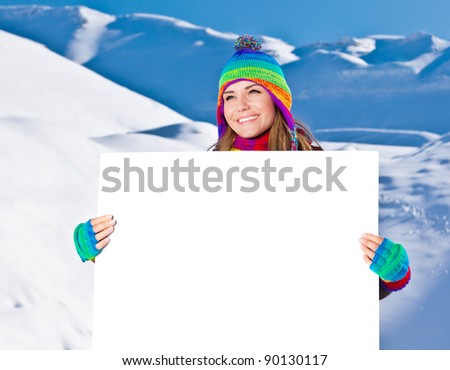 Happy girl holding white board, winter outdoor portrait, beautiful female teen in colorful hat, young pretty smiling woman holding blank paper card in hands, text space, copyspace