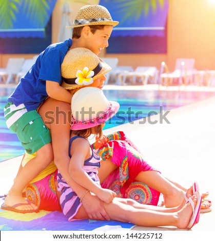 Happy family sitting near poolside, mother with two cute kids enjoying summer holidays, little sister and brother with young mom on luxury resort, love concept