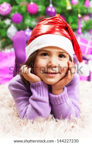 Closeup portrait of cute little arabic girl wearing red Santa Claus hat, lying down on the floor near beautiful decorated Christmas tree, New Year party