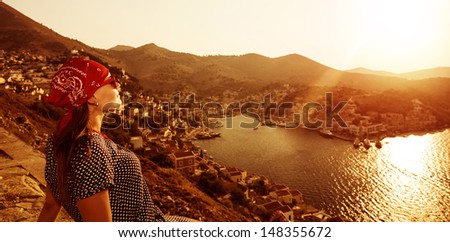 Tourist girl sitting and looking to the sea, beautiful orange sunset, coastal city, mountains landscape, traveling and tourism concept