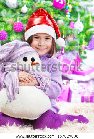 Cute little girl sitting on the floor under big decorated Christmas tree, wearing red Santa hat, holding in hands white snowman soft toy, New Year holidays