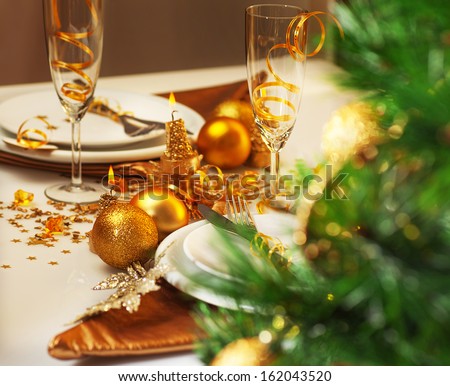 Photo Of Luxury Christmastime Table Setting, Holiday Dinner In Restaurant, Festive White Dinnerware Decorated With Pretty Golden Balls And Ribbons, Warm Candle Light, Green Christmas Tree In Room