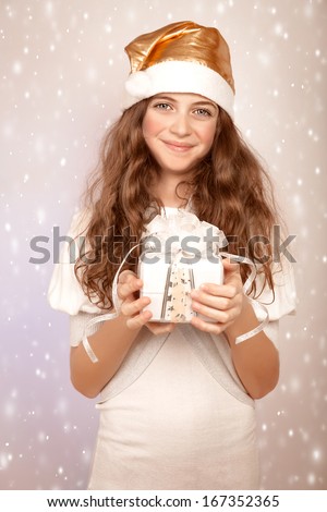 Sweet Santa helper, pretty teen girl wearing festive hat and holding in hands gift box, snow fall, Christmas party concept