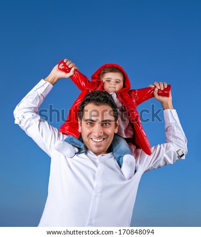 Portrait of cheerful young father with cute little son sitting on his shoulders on blue clear shy background, having fun outdoors