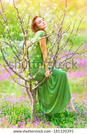 Beautiful forest nymph wearing long gentle green dress, sitting on the tree, blooming nature, romantic fairytale, spring season concept