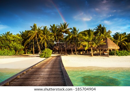 Beautiful exotic beach landscape, wooden pier on clear turquoise water, colorful rainbow over luxury island resort, summer vacation and holidays concept