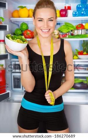 Healthy woman with fresh salad standing near open fridge full of vegetables, athletic girl with measure tape, perfect body, organic food concept