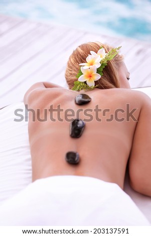 Enjoying day spa, cute female with frangipani flowers in hair lying down on massage table on the beach, black hot stones therapy, zen balance concept