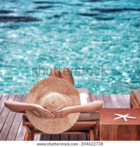 Young woman sitting on wooden pier on the sea wearing hat and taking sun bath, enjoying perfect summer day, traveling and luxury vacation concept