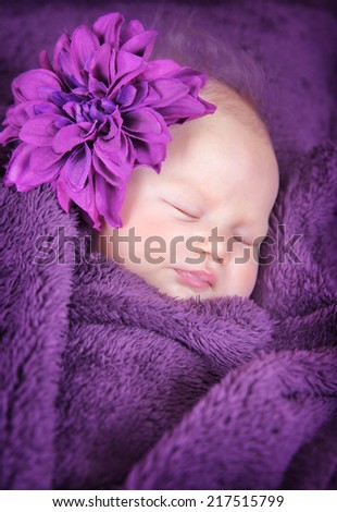 Closeup portrait of cute little newborn baby sleeping at home covered in purple blanket and wearing big stylish flower accessories, innocence concept