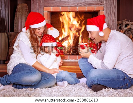 Happy family by fireplace, parents with baby girl wearing Santa Claus hats, sitting relaxed in ski resort chalet and drinking hot tea, joyful winter vacation, people traveling on Christmas holidays