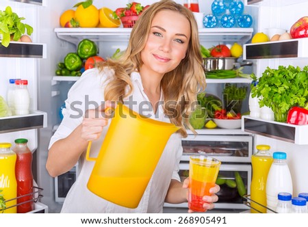 Happy woman standing near open refrigerator full of fresh fruits and vegetables and pouring juice in glass, healthy eating concept