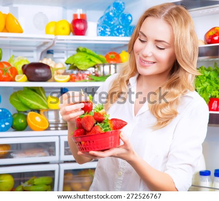 Portrait of a beautiful female standing near open fridge full of fruits and vegetables, eating fresh red ripe juicy strawberries, enjoying healthy nutrition, wellness and healthy living