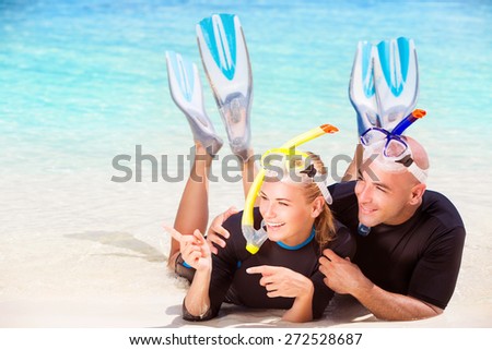 Joyful diver couple lying down on the beach, wearing mask and flippers for snorkeling, looking on the side and showing on something, active summer time vacation