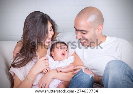 Portrait of happy cheerful family sitting on the couch at home, young parents enjoying time spent with their adorable newborn daughter