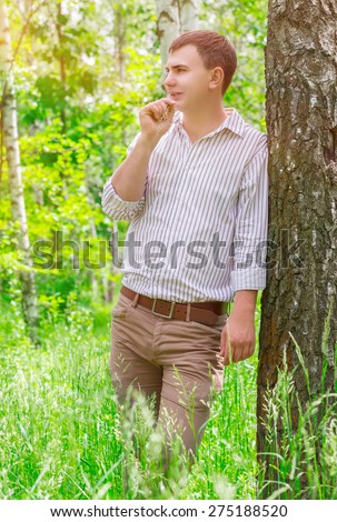 Handsome guy in spring park, standing near birch tree with straw in mouth and dreamy looking in side, spending warm sunny day outdoors