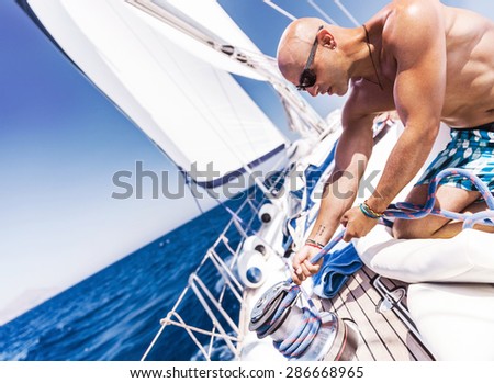 Handsome shirtless sailor working on sailboat, pulling rope on crank, summer time activity, spending vacation in a sea traveling