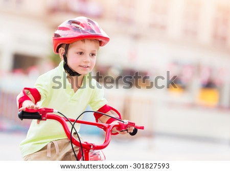 Little boy playing outdoors, cute cheerful child having fun in summer camp, nice kid riding on red stylish bicycle on blur urban background, enjoying carefree summer holidays