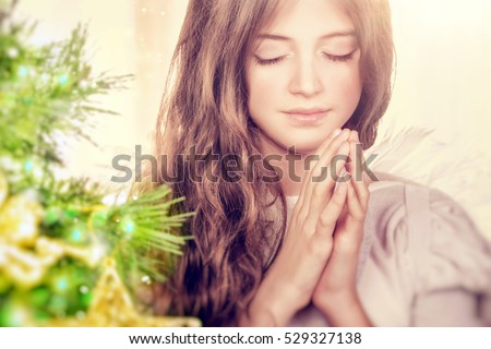 Closeup portrait of a beautiful calm girl with closed eyes praying near Christmas tree, gentle young angel wishing peace and harmony for everyone, happy religious holiday