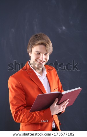 Angry young teacher   / Portrait of a young, handsome man in the orange jacket standing on black background holding blackboard book