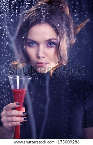Woman with a glass of red wine . Beautiful mysterious woman dressed in black dress looking out the window holding in his hand a glass of red wine .