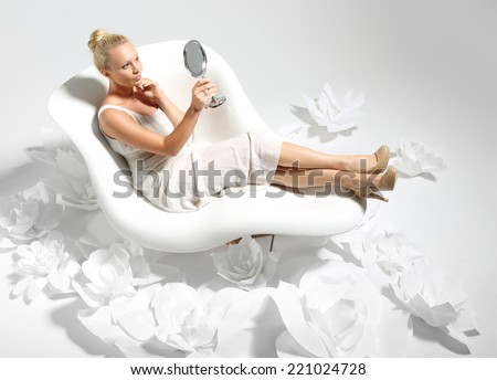 Mirror, mirror, girl looks in the mirror .Beautiful young woman sitting on a white chair surrounded by stylish white flowers