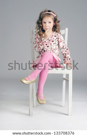Fashionable little cute girl sitting thoughtful on the white chair, posing, looking away.
