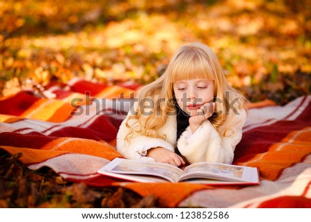 Cute little girl with curly blond hair lies on the bright plaid and reads a book  in autumn park on a sunny day