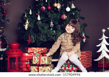 Girl With Dark Hair Sitting On A Box With Gifts. Christmas Tree In The Background. Smiles