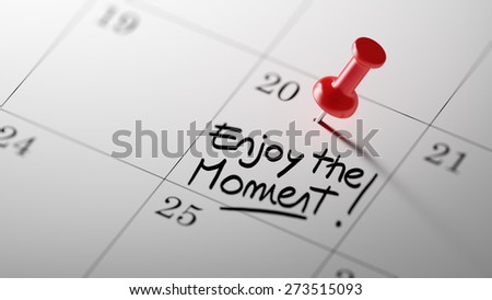 Concept image of a Calendar with a red push pin. Closeup shot of a thumbtack attached. The words Enjoy the moment written on a white notebook to remind you an important appointment.
