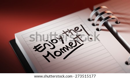 Closeup of a personal agenda setting an important date representing a time schedule. The words Enjoy the moment written on a white notebook to remind you an important appointment.
